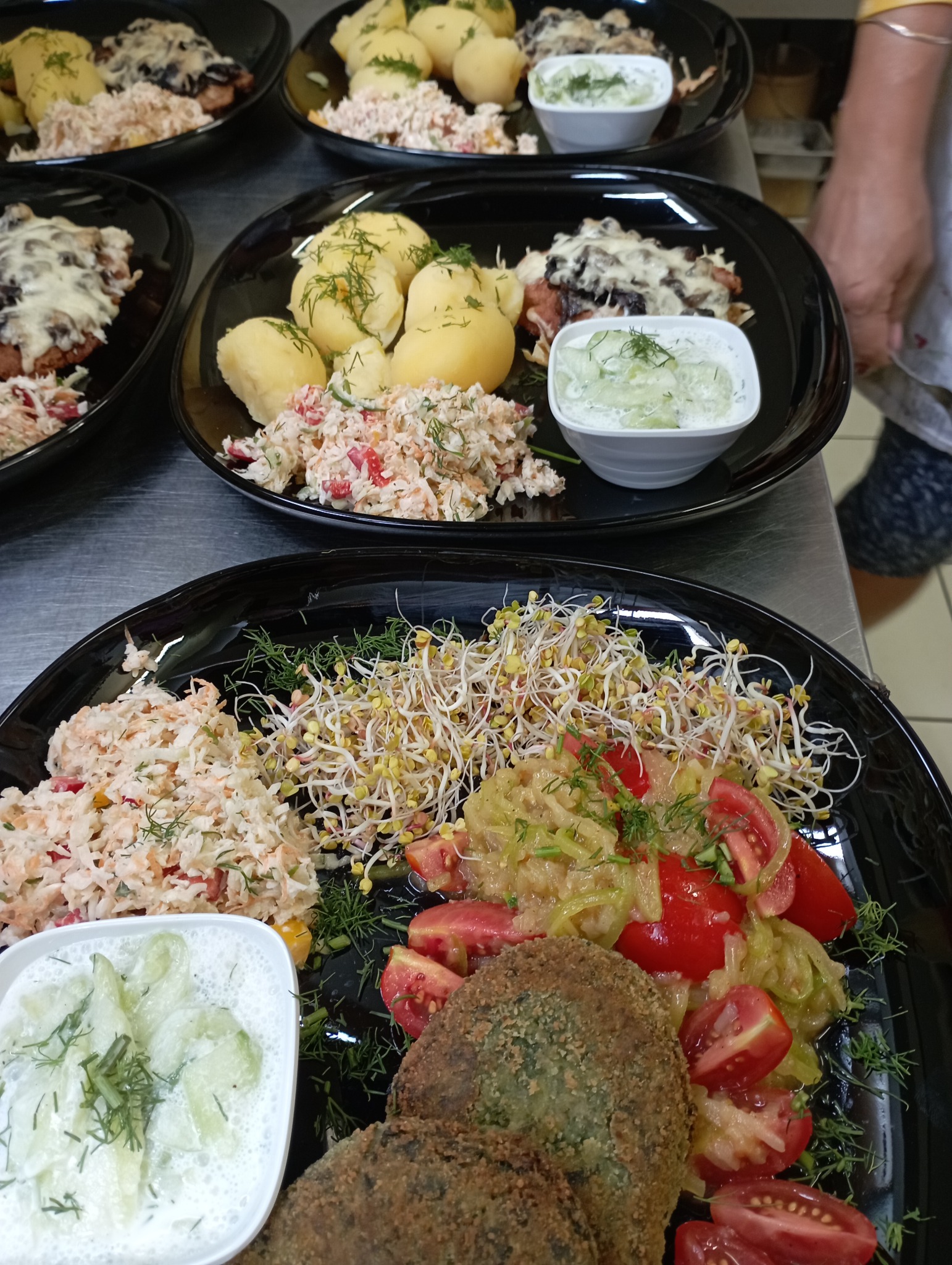 Czarne talerze z gotowymi daniami: kotlety w panierce, ziemniaki posypane koperkiem, sałatka colesław, sałatka ze świeżych ogórków w śmietanie, kiełki, pomidory i duszona kapusta.