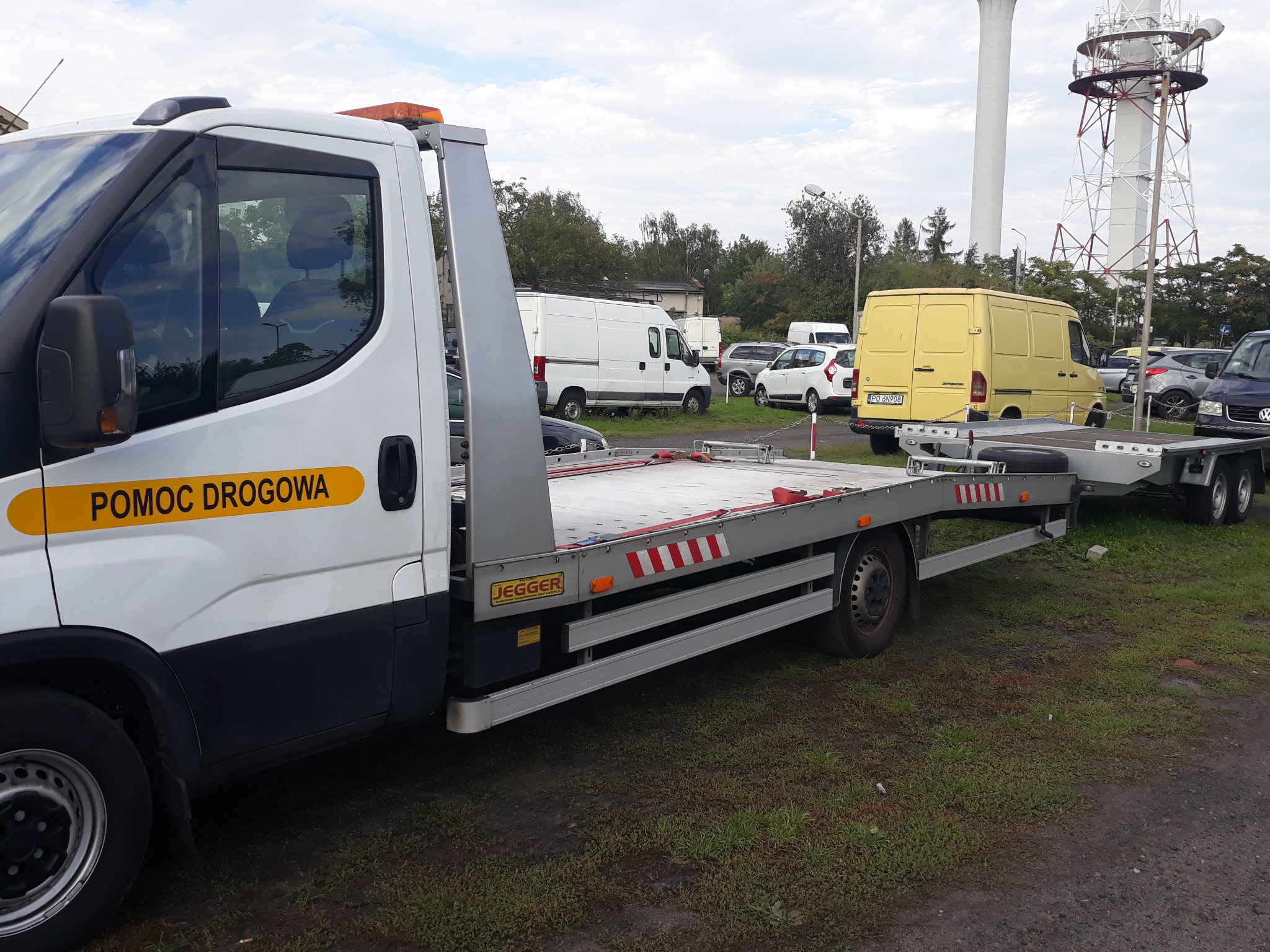 Biała laweta z napisem 'POMOC DROGOWA' na żółtym tle, zaparkowana na placu z widoczną wieżą telekomunikacyjną w tle.