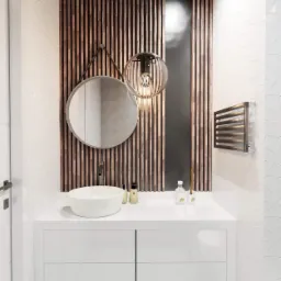 Nowoczesna łazienka z umywalką nablatową, okrągłym lustrem na pasku i dekoracyjną lampą wiszącą na tle ściany z drewnianych listew i geometrycznych płytek.