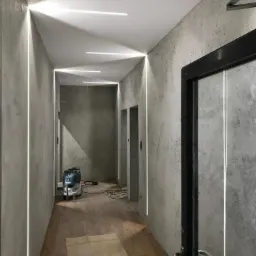 Korytarz z oświetleniem LED wpuszczonym w ściany, surowe szare ściany z efektem betonu, drewniana podłoga częściowo zakryta kartonem, widoczny odkurzacz przemysłowy i czarna rama drzwi z metalową...