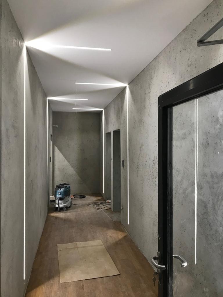 Korytarz z oświetleniem LED wpuszczonym w ściany, surowe szare ściany z efektem betonu, drewniana podłoga częściowo zakryta kartonem, widoczny odkurzacz przemysłowy i czarna rama drzwi z metalową...