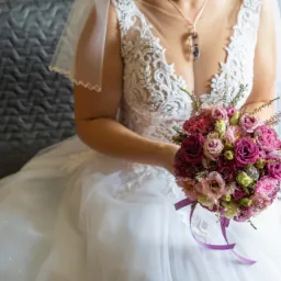 Panna młoda w koronkowej sukni ślubnej trzyma bukiet z różowych i fioletowych róż, widoczny wisiorek z suszonymi kwiatami na szyi, szary pikowany zagłówek w tle.