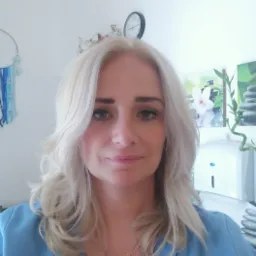 Portret kobiety z blond włosami w niebieskim uniformie, na szyi złoty medalik, w tle gabinet z dekoracjami i zegarem.