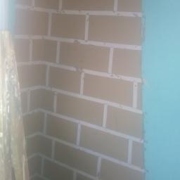 Pb-Bud - Ściana z brązowych płyt kartonowo-gipsowych imitujących cegłę, z widocznymi spoinami wypełnionymi szpachlą, w rogu pomieszczenia, obok fragment niebieskiej ściany i drabiny.