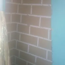 Ściana z brązowych płyt kartonowo-gipsowych imitujących cegłę, z widocznymi spoinami wypełnionymi szpachlą, w rogu pomieszczenia, obok fragment niebieskiej ściany i drabiny.