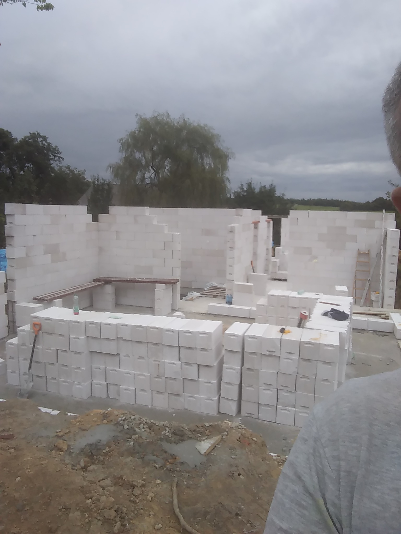 Budowa domu z białych bloczków, widoczne wznoszone ściany, palety z bloczkami, drabina i narzędzia, pochmurne niebo w tle.