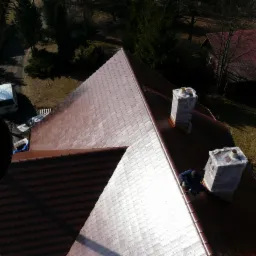 Widok z góry na fragment dachu z brązową dachówką, z dwoma kominami zabezpieczonymi folią, pracownik w niebieskim kombinezonie przy krawędzi, biały samochód dostawczy w tle.