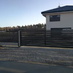 Nowoczesne, czarne ogrodzenie metalowe z poziomymi panelami i perforowanymi wzorami na tle domu z ciemnym dachem.