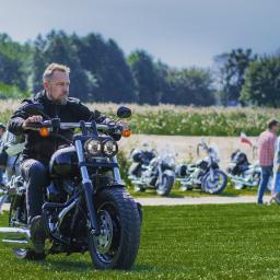 Piter_fotografuje - Mężczyzna z brodą na czarnym motocyklu Harley-Davidson, w tle inne motocykle i osoby na pikniku charytatywnym.