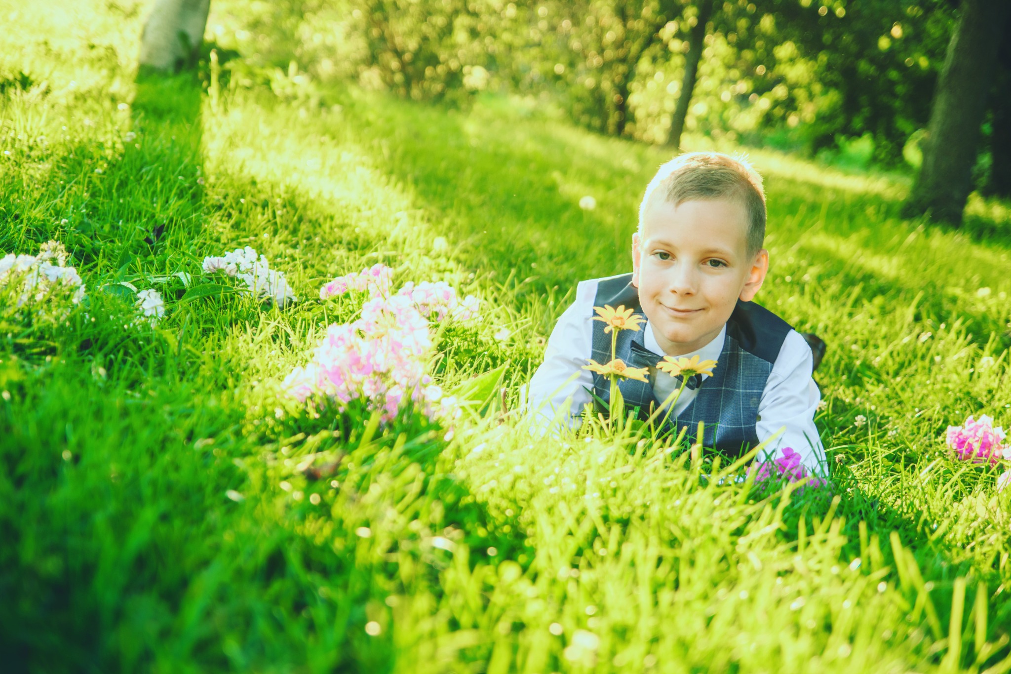 Uśmiechnięty chłopiec w stroju komunijnym leży na trawie wśród kwiatów, z delikatnym bokeh w tle.