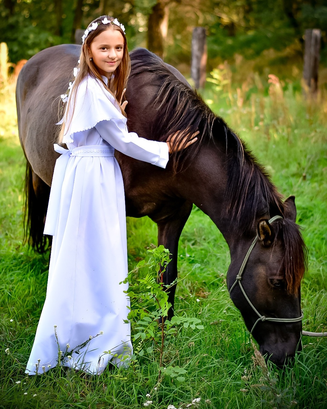 Dziewczynka w białej sukience komunijnej z wiankiem na głowie, delikatnie głaszcze ciemnego konia na zielonej łące w słoneczny dzień.
