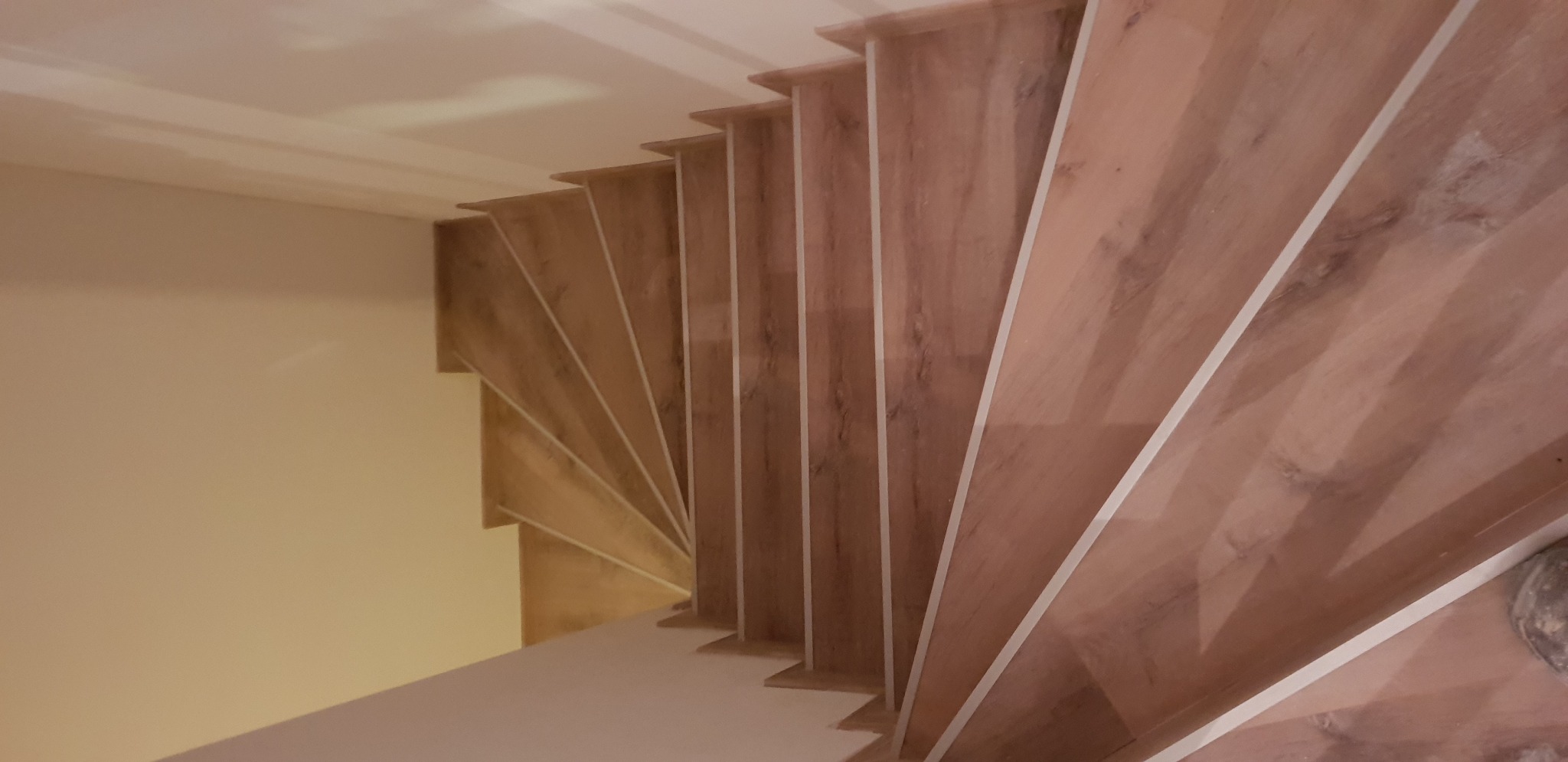 Widok z góry na drewniane schody z białymi listwami, skręcające w lewo, oświetlone ciepłym światłem.