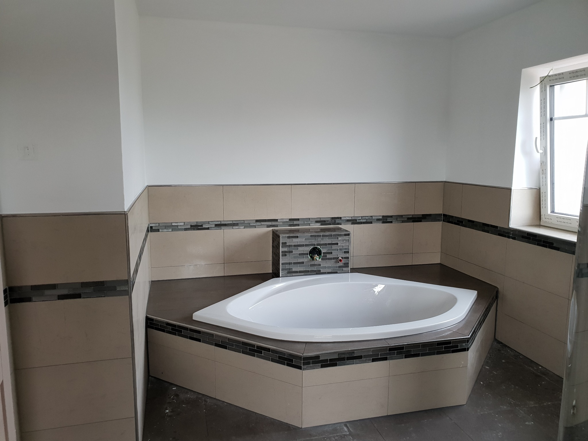 Narożna wanna w obudowie z płytek w trakcie wykańczania łazienki, widoczna ściana z przygotowanymi przyłączami do baterii, jasne płytki i mozaika, okno z białymi ramami.