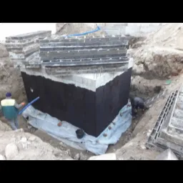 Betonowy fundament szamba zabezpieczony czarną izolacją bitumiczną, widoczni dwaj pracownicy podczas prac ziemnych, szalunki w tle.