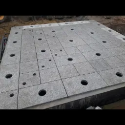 Szara, granitowa powierzchnia składająca się z kwadratowych płyt z wywierconymi otworami o różnych średnicach, widoczna na placu budowy w pochmurny dzień.