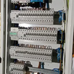 Rozdzielnica elektryczna z zainstalowanymi zabezpieczeniami i okablowaniem, widoczny licznik energii elektrycznej oraz listwy zaciskowe, tło stanowi surowa ściana z widocznymi uszkodzeniami.