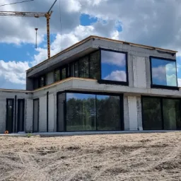 Surowy stan nowoczesnego domu z dużymi, czarnymi ramami okiennymi, widok z zewnątrz na plac budowy z piaskiem i żurawiem budowlanym w tle.