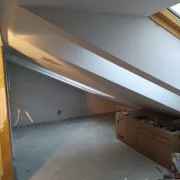 Poddasze po remoncie z widocznymi skośnymi ścianami pomalowanymi na szaro, okno dachowe w drewnianej ramie. Widoczna puszka farby i kartonowe pudła. Ściany świeżo pomalowane, brak dekoracji.