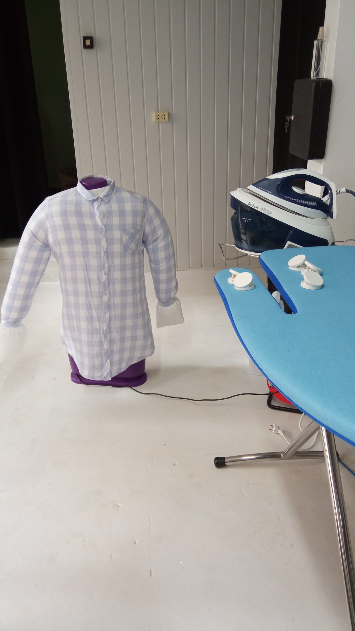 Manekin parowy do prasowania koszul w niebieską kratę obok deski do prasowania i żelazka parowego Tefal.