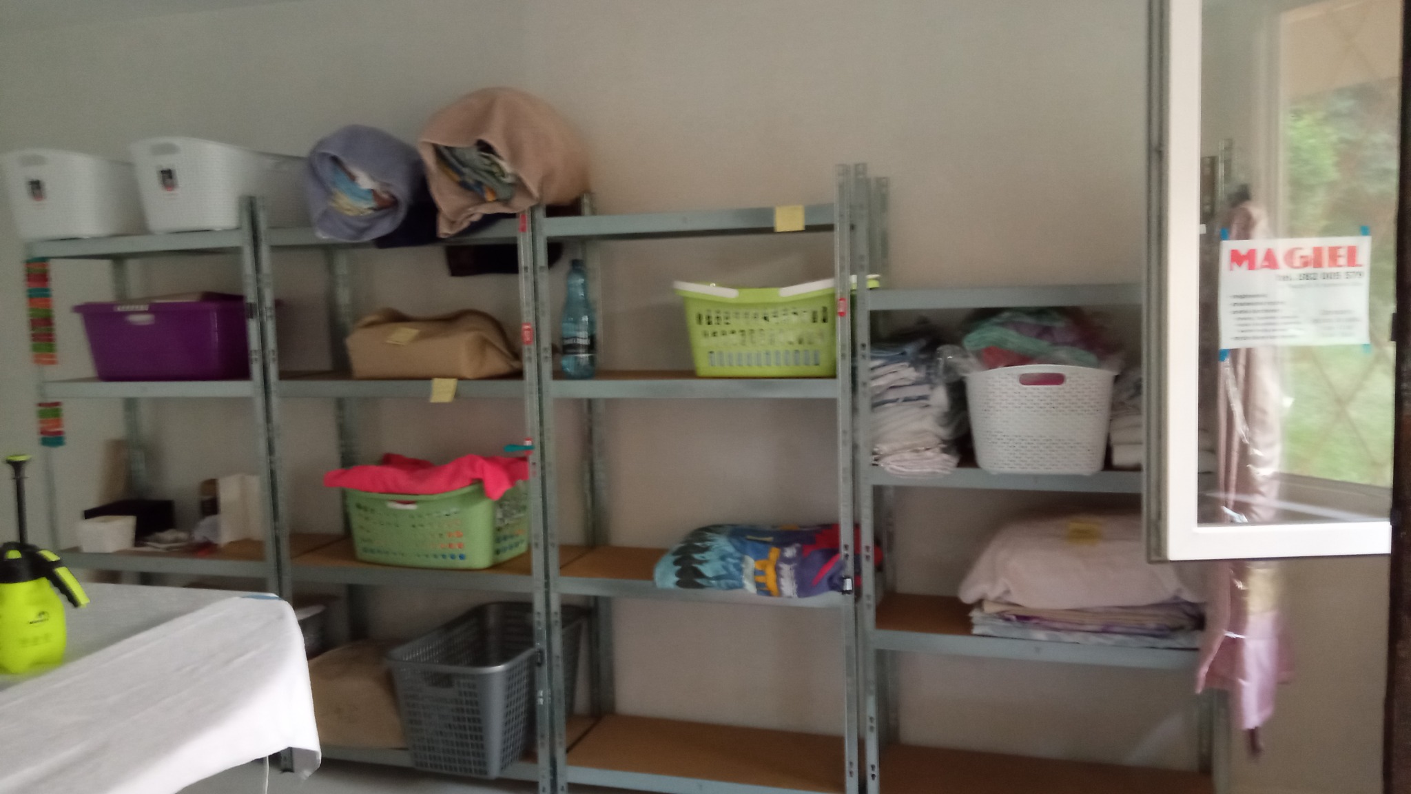 Metalowe regały z koszami i złożonymi tekstyliami w pomieszczeniu pralni, widoczny spryskiwacz i ulotka firmy na szybie okna.