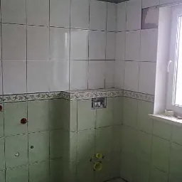 Ściany łazienki w trakcie układania płytek, widoczne przygotowane otwory na instalacje, dekoracyjny pas płytek na styku dwóch kolorów, oraz okno z parapetem.