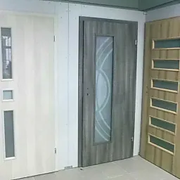 Ekspozycja trzech różnych modeli drzwi z przeszkleniami: białe drzwi z trzema małymi szybkami, szare drzwi z dużą szybą z geometrycznym wzorem oraz drzwi w odcieniach drewna z pięcioma poziomymi...