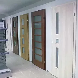 Ekspozycja różnorodnych skrzydeł drzwiowych z przeszkleniami, ustawionych w rzędzie wzdłuż ściany w pomieszczeniu sklepowym.
