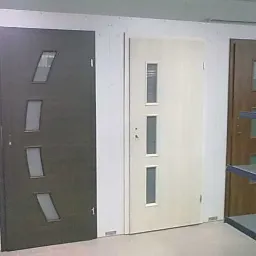 Ekspozycja trzech różnych modeli drzwi z szybami: ciemne drzwi z trzema ukośnymi szybami, jasne drzwi z trzema pionowymi szybami i brązowe drzwi z dużą szybą w górnej części.