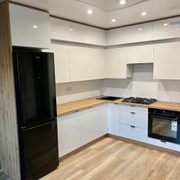 Juliusz Bieszczanin - Nowoczesna kuchnia na wymiar z białymi, lakierowanymi frontami, drewnianymi blatami i czarnym sprzętem AGD, w tym lodówką i piekarnikiem, na tle zielonej ściany.