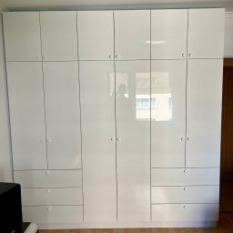 Juliusz Bieszczanin - Biała, błyszcząca zabudowa meblowa z szafkami i szufladami, z okrągłymi uchwytami, odbijająca w lustrzanych frontach fragment budynku za oknem.