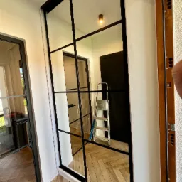 Szklana ściana działowa z czarnymi szprosami, odbijająca wnętrze z drabinką i parkietem w jodełkę, widoczna podczas montażu.