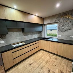 Juliusz Bieszczanin - Jasne drewniane szafki kuchenne z czarnymi blatami i górnymi szafkami, podświetlone, z wzorem mandali na ścianie i drewnianą podłogą w jodełkę, okno z widokiem na dom z panelami słonecznymi.