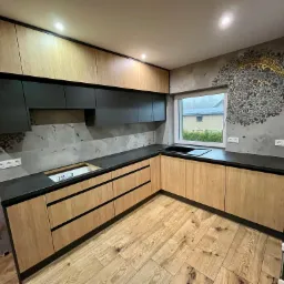 Jasne drewniane szafki kuchenne z czarnymi blatami i górnymi szafkami, podświetlone, z wzorem mandali na ścianie i drewnianą podłogą w jodełkę, okno z widokiem na dom z panelami słonecznymi.