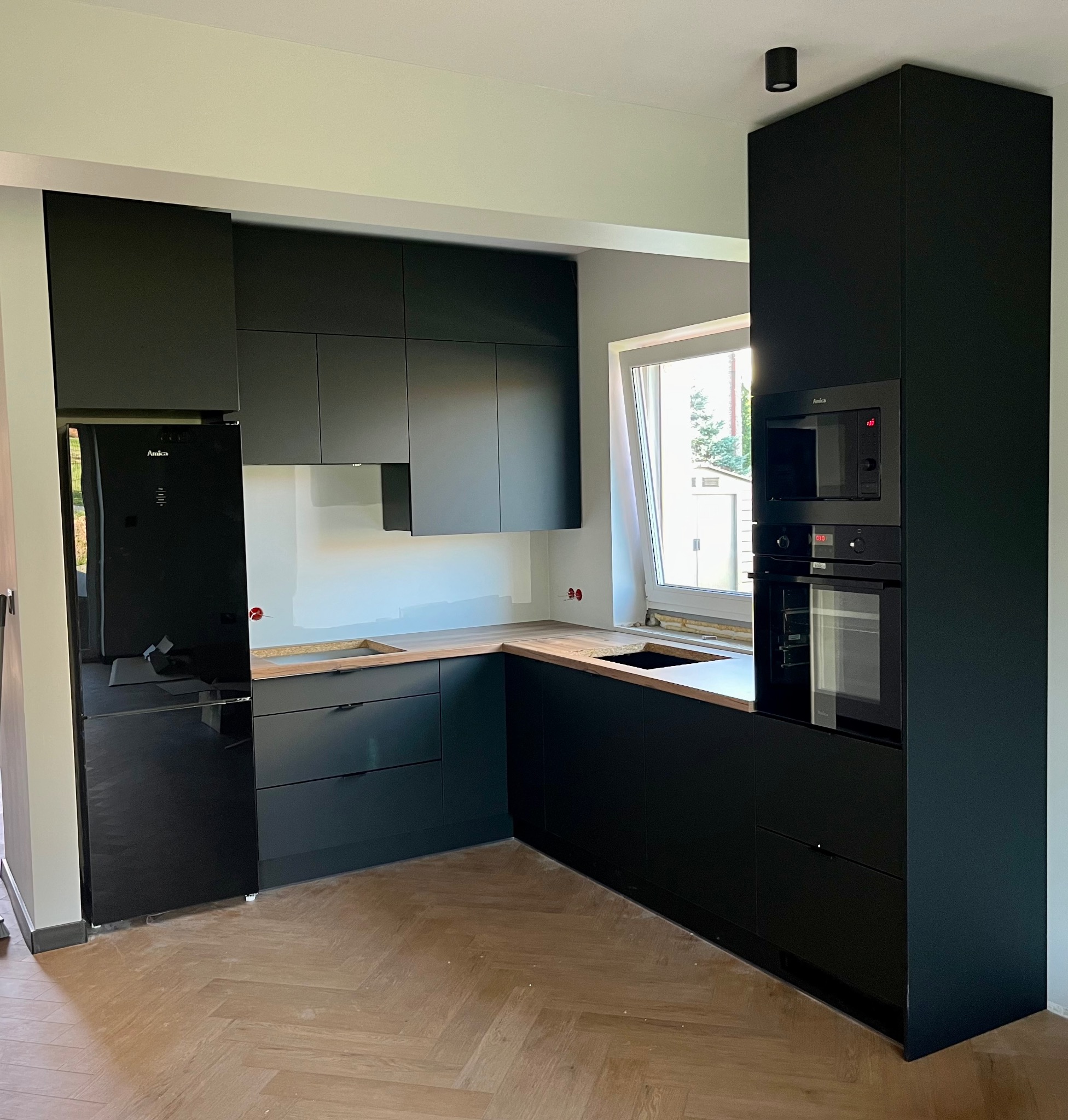 Nowoczesna, czarna zabudowa kuchenna z drewnianym blatem, z widoczną lodówką Amica i wbudowanym piekarnikiem oraz mikrofalówką, na tle jasnych ścian i podłogi w jodełkę.