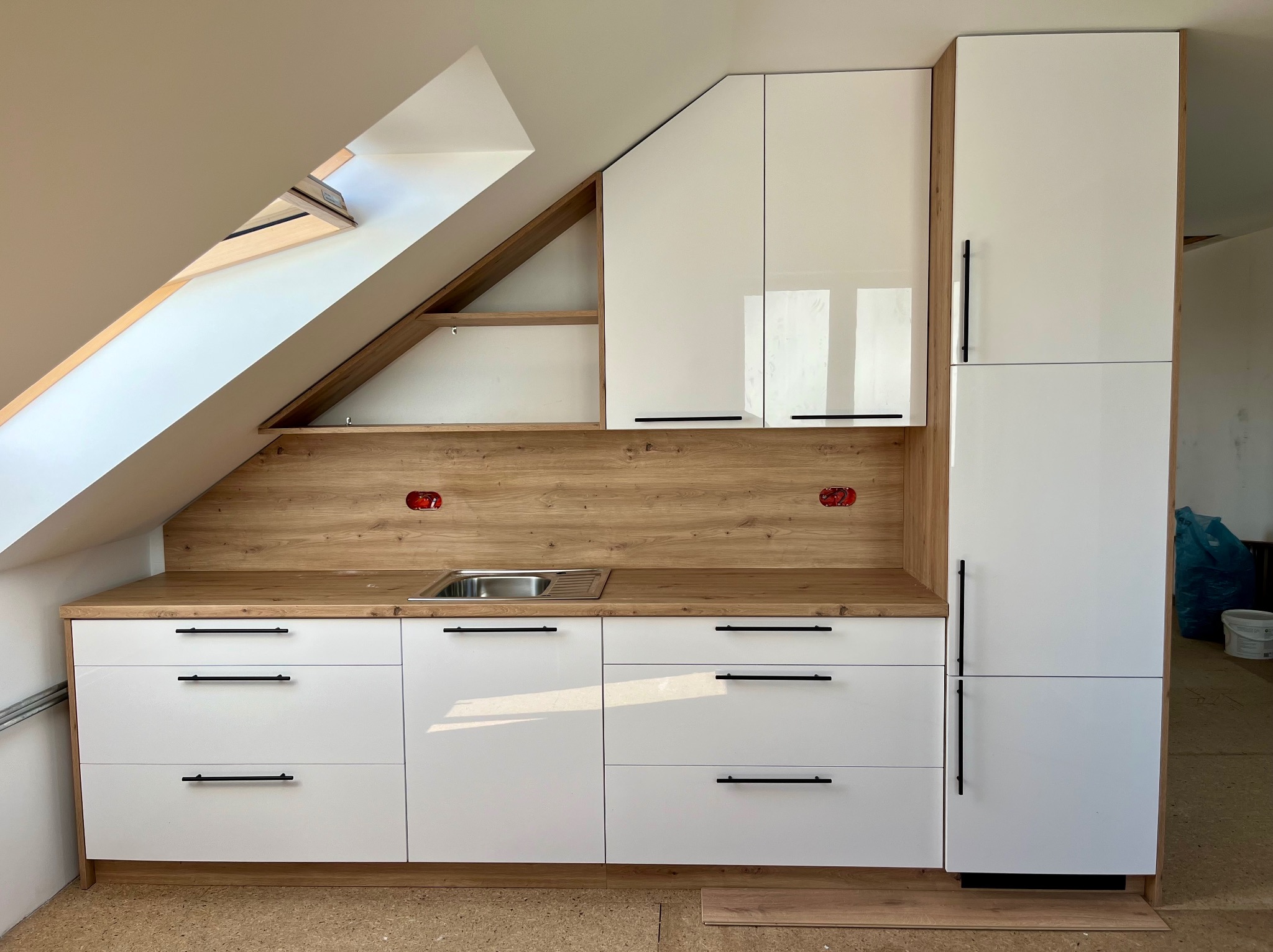 Biała kuchnia z drewnianymi akcentami pod skosem dachu z oknem dachowym i odsłoniętymi gniazdkami elektrycznymi.