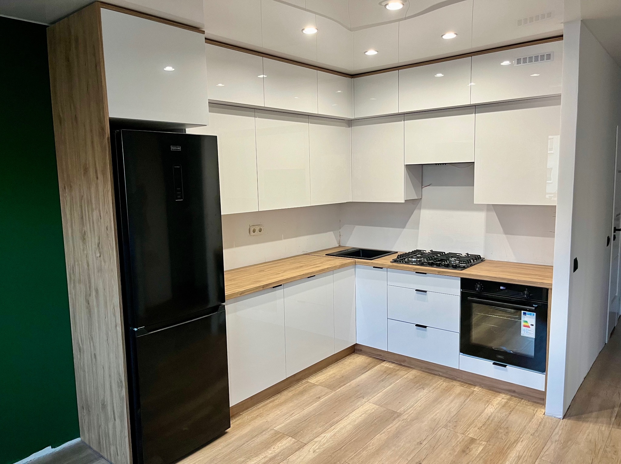 Nowoczesna kuchnia na wymiar z białymi, lakierowanymi frontami, drewnianymi blatami i czarnym sprzętem AGD, w tym lodówką i piekarnikiem, na tle zielonej ściany.
