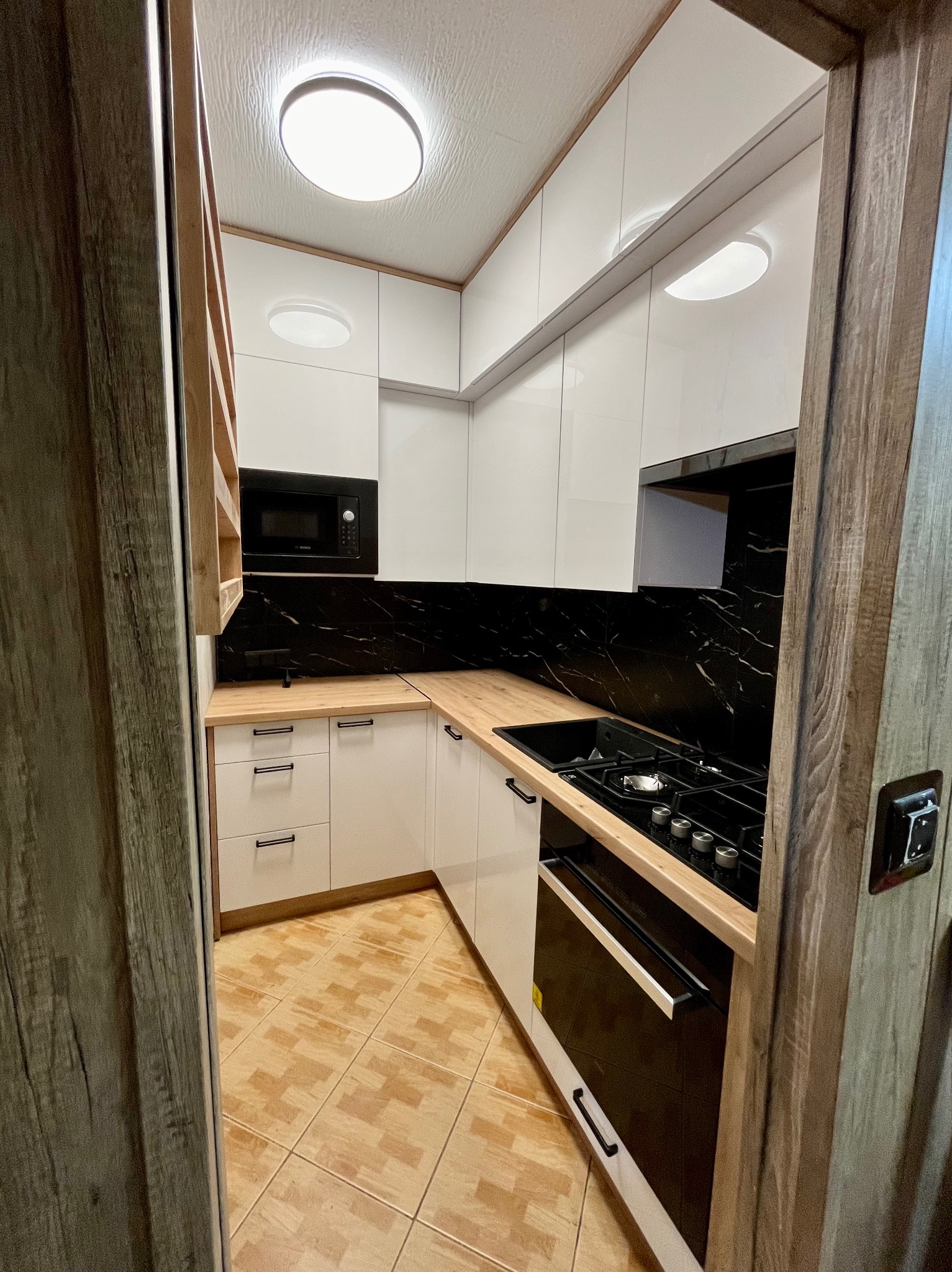 Nowoczesna kuchnia z białymi szafkami, czarnym blatem imitującym marmur i drewnianymi blatami, widziana zza drewnianej framugi drzwi.