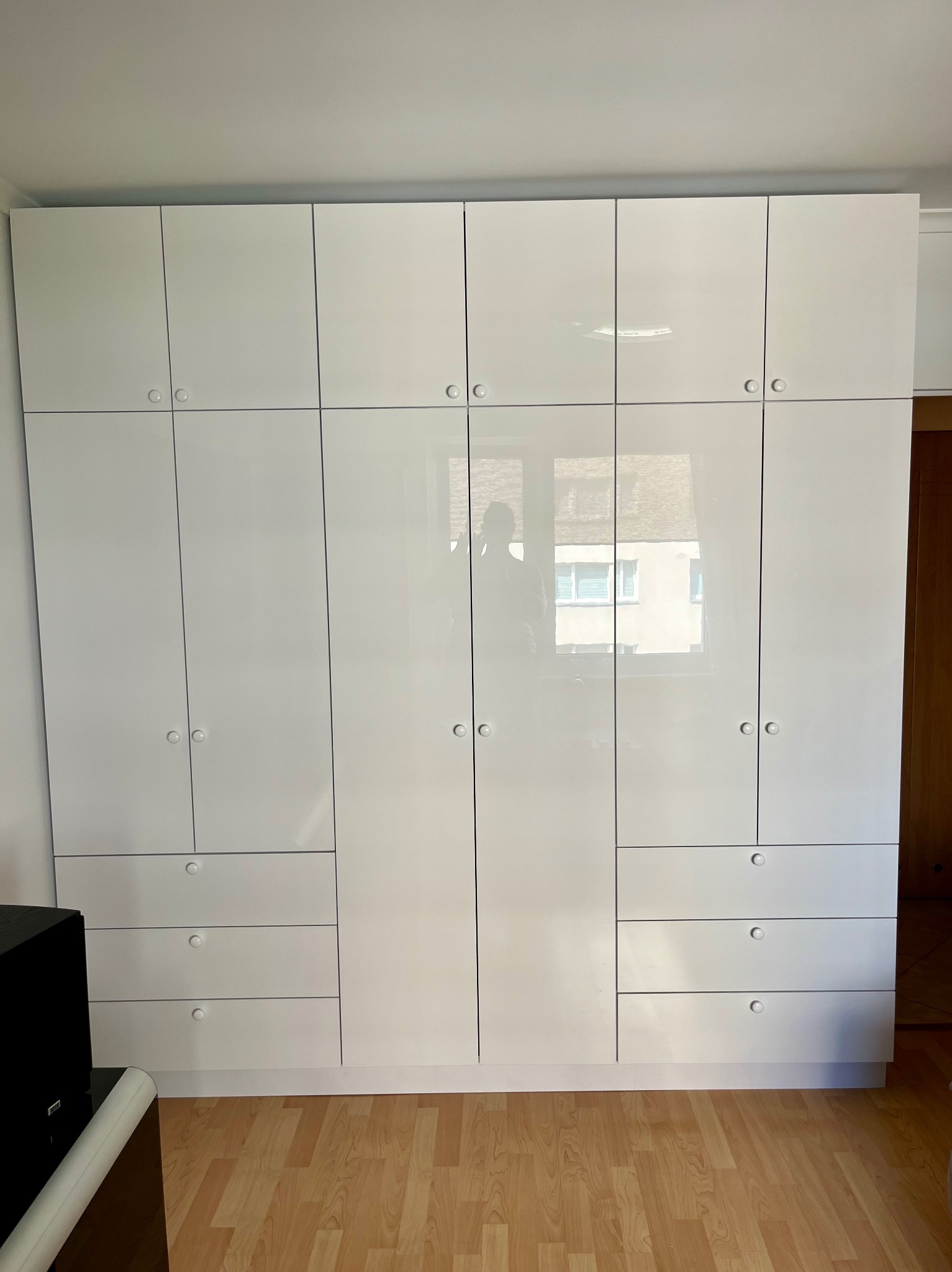 Biała, błyszcząca zabudowa meblowa z szafkami i szufladami, z okrągłymi uchwytami, odbijająca w lustrzanych frontach fragment budynku za oknem.