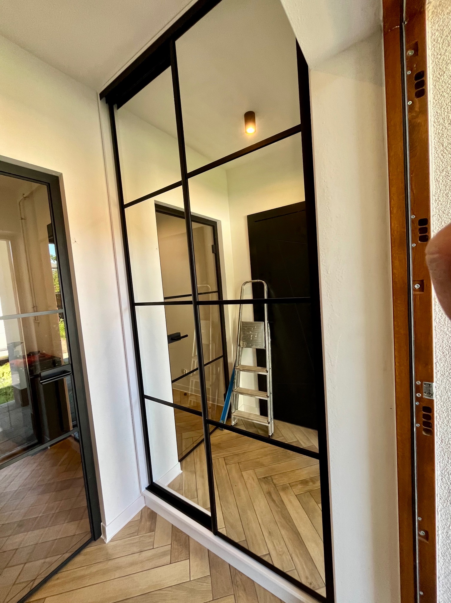 Szklana ściana działowa z czarnymi szprosami, odbijająca wnętrze z drabinką i parkietem w jodełkę, widoczna podczas montażu.