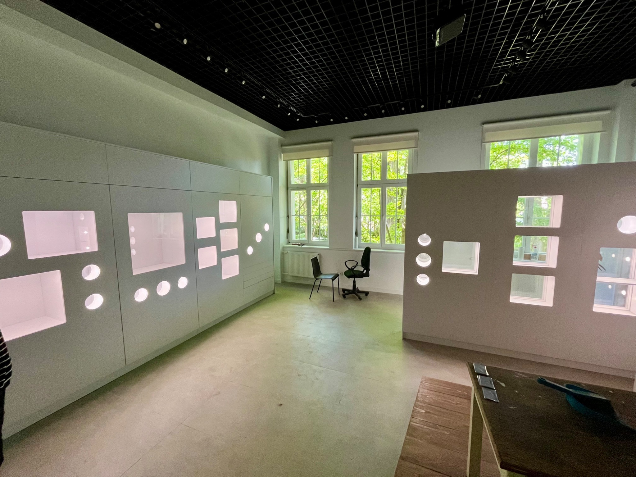 Jasne pomieszczenie z ekspozycją meblową: białe szafy z podświetlanymi wnękami o różnych kształtach, widoczne krzesła biurowe i fragment stołu z narzędziami, sufit z czarną kratownicą.