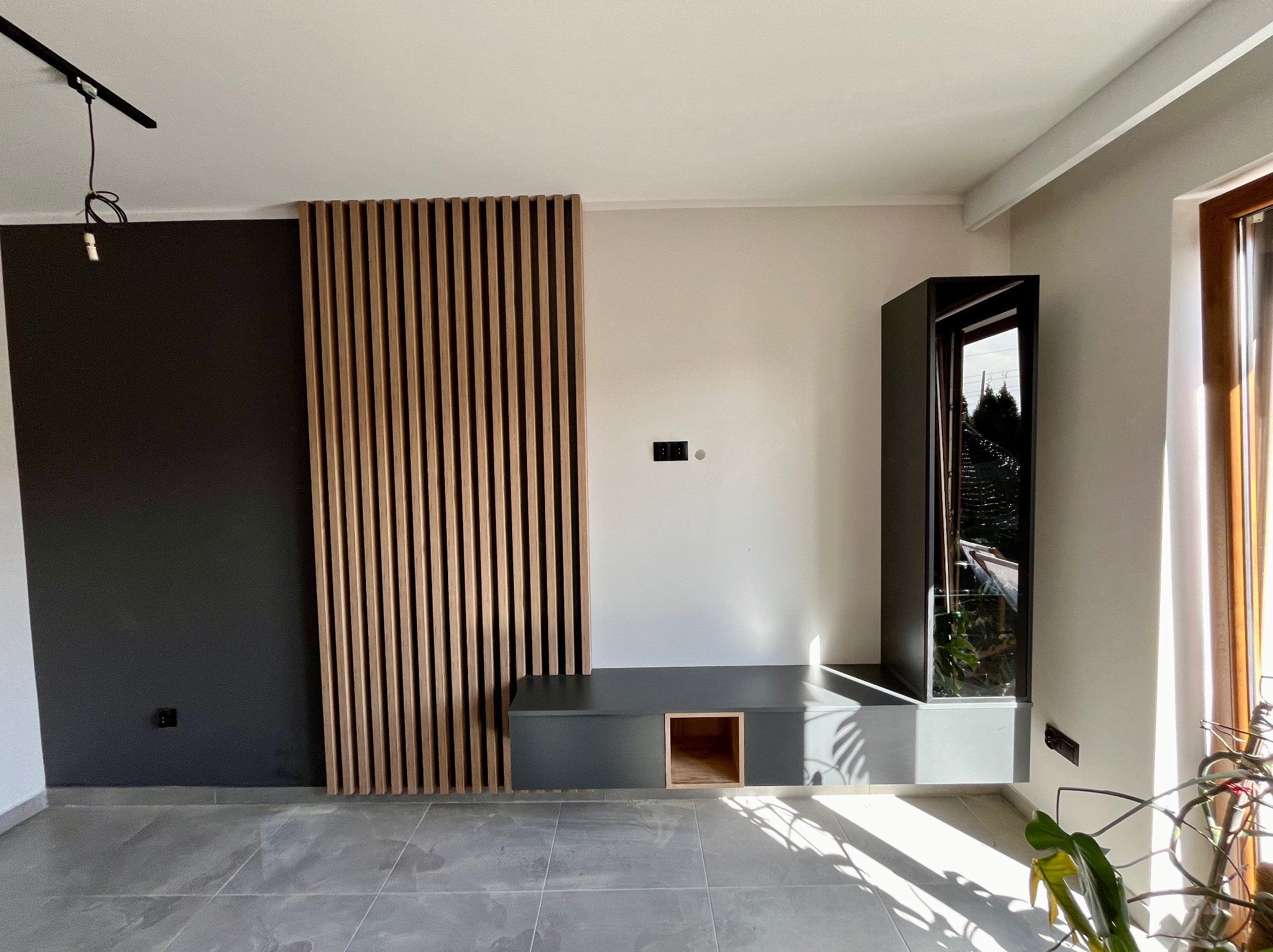 Nowoczesny salon z szarą zabudową meblową, drewnianymi lamelami na ścianie i wysoką witryną z odbijającym się widokiem na zewnątrz, oświetlony naturalnym światłem padającym przez okno.