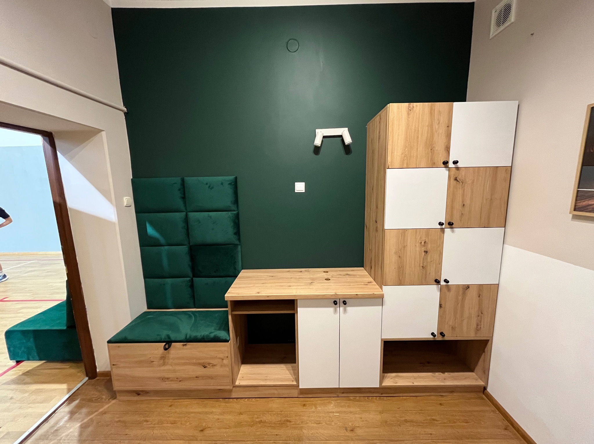 Zabudowa wnęki z szafkami z frontami drewnopodobnymi i białymi, siedziskiem z zielonym obiciem i panelem ściennym z zielonych kwadratów.