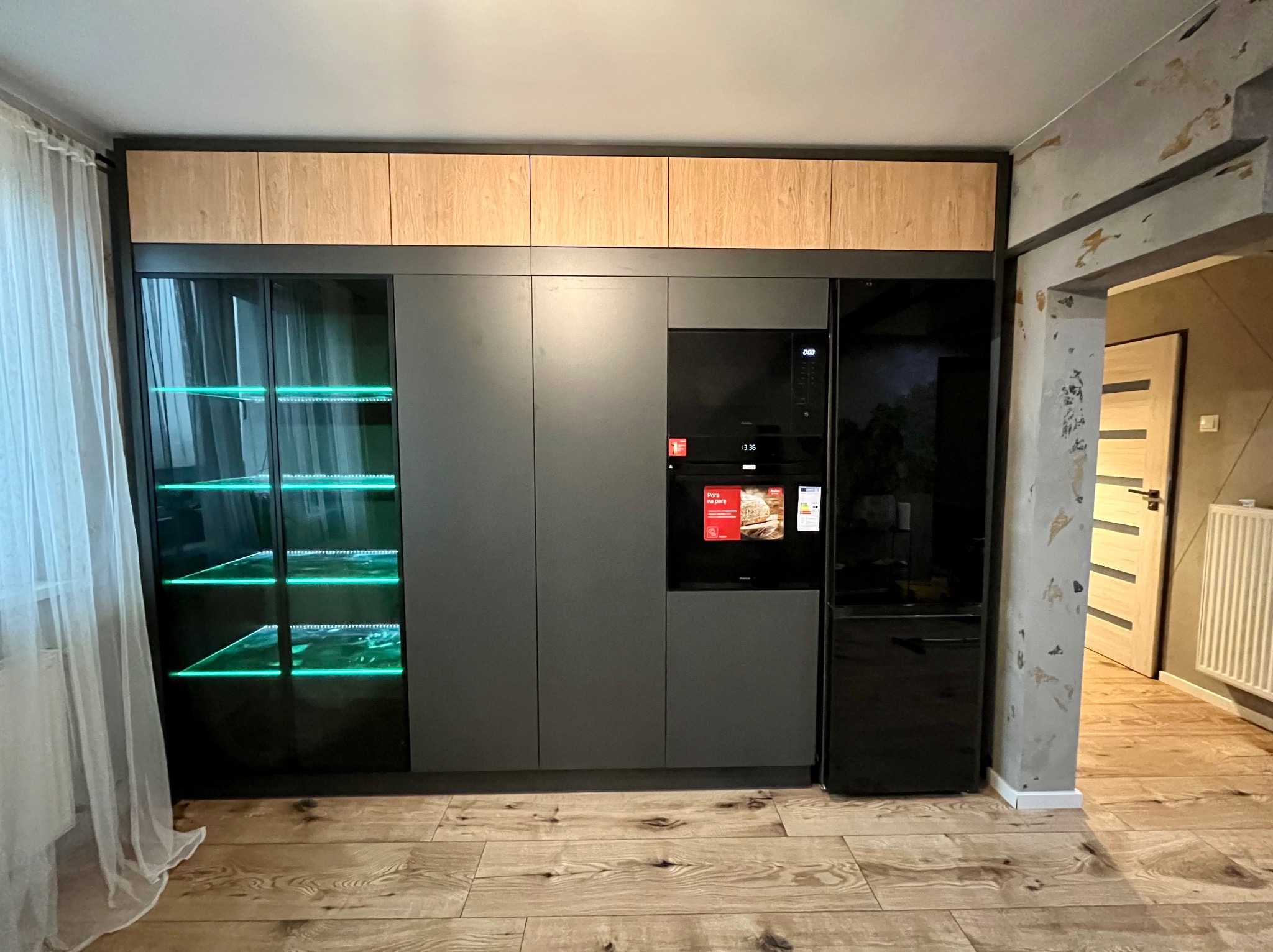 Nowoczesna zabudowa kuchenna w szarościach z drewnianymi akcentami, oświetlona szafka z zielonym podświetleniem LED, sprzęt AGD w zabudowie, parkiet na podłodze.