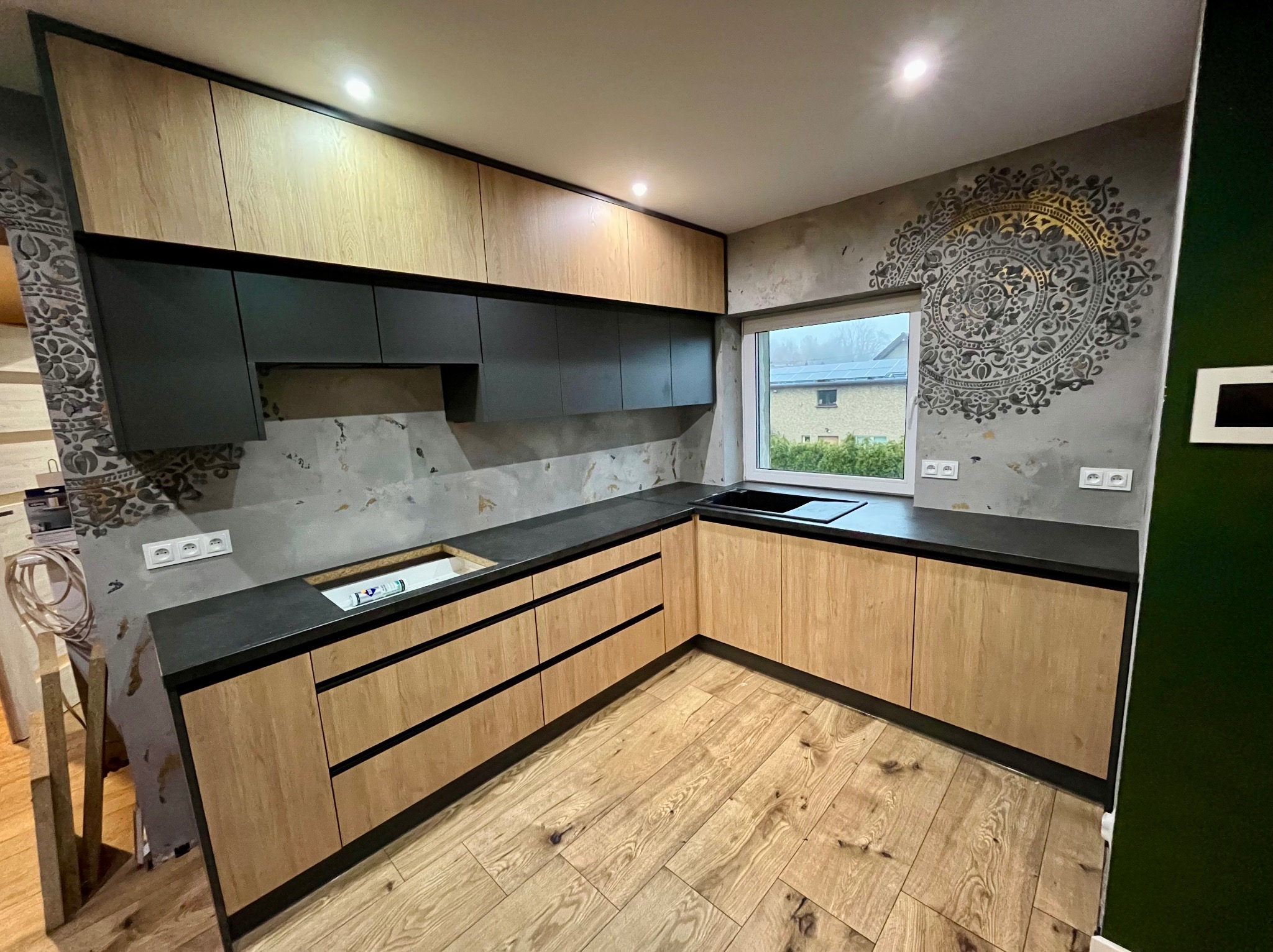 Jasne drewniane szafki kuchenne z czarnymi blatami i górnymi szafkami, podświetlone, z wzorem mandali na ścianie i drewnianą podłogą w jodełkę, okno z widokiem na dom z panelami słonecznymi.