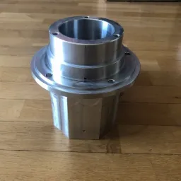 Metalowy element z otworem i kołnierzem, obrobiony na kształt wielokąta, leżący na parkiecie.