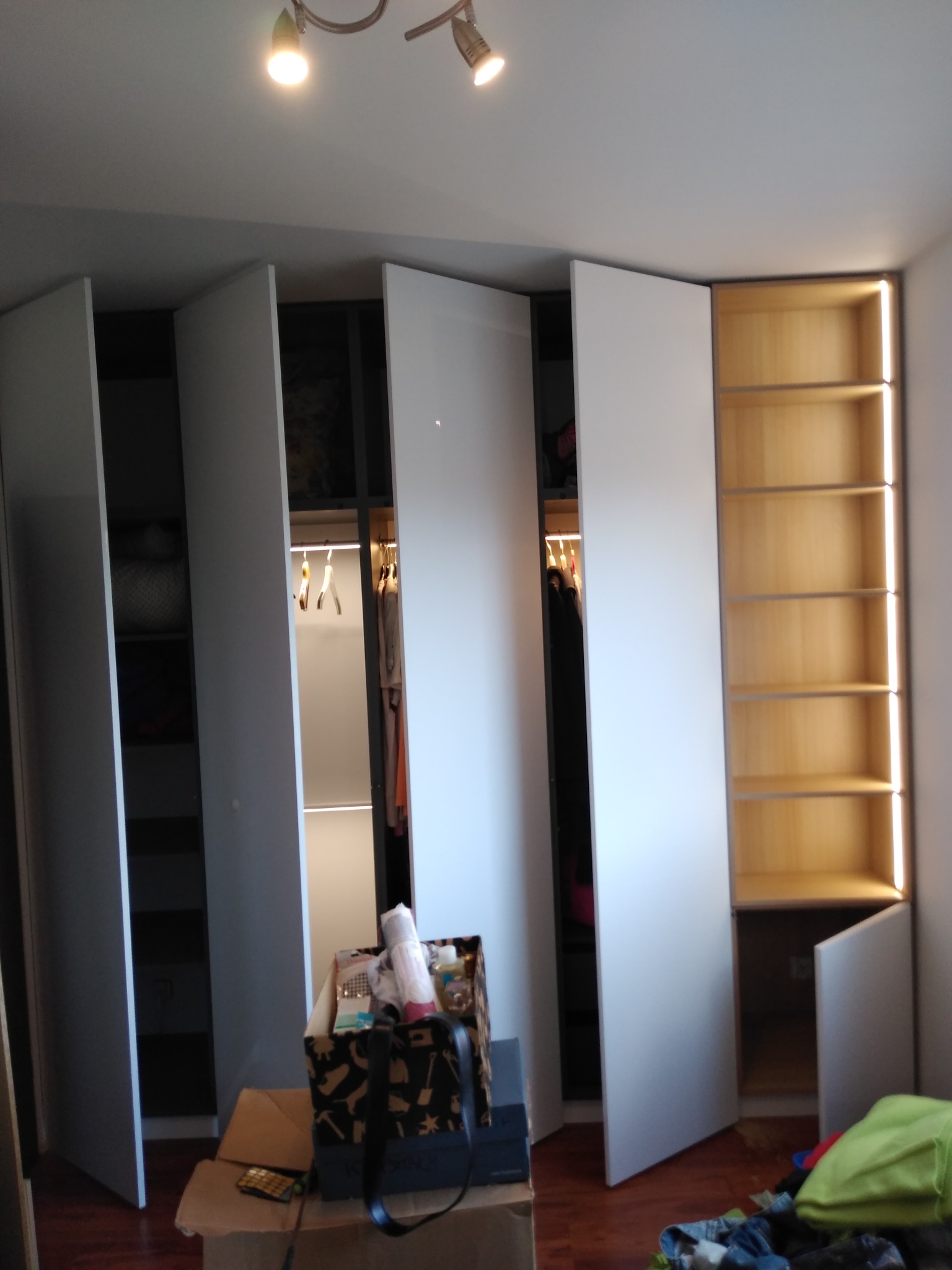 Nowoczesna szafa z białymi frontami, oświetleniem LED wewnątrz, częściowo otwarta, z widocznymi ubraniami na wieszakach i półkami, kartony i rzeczy na podłodze.