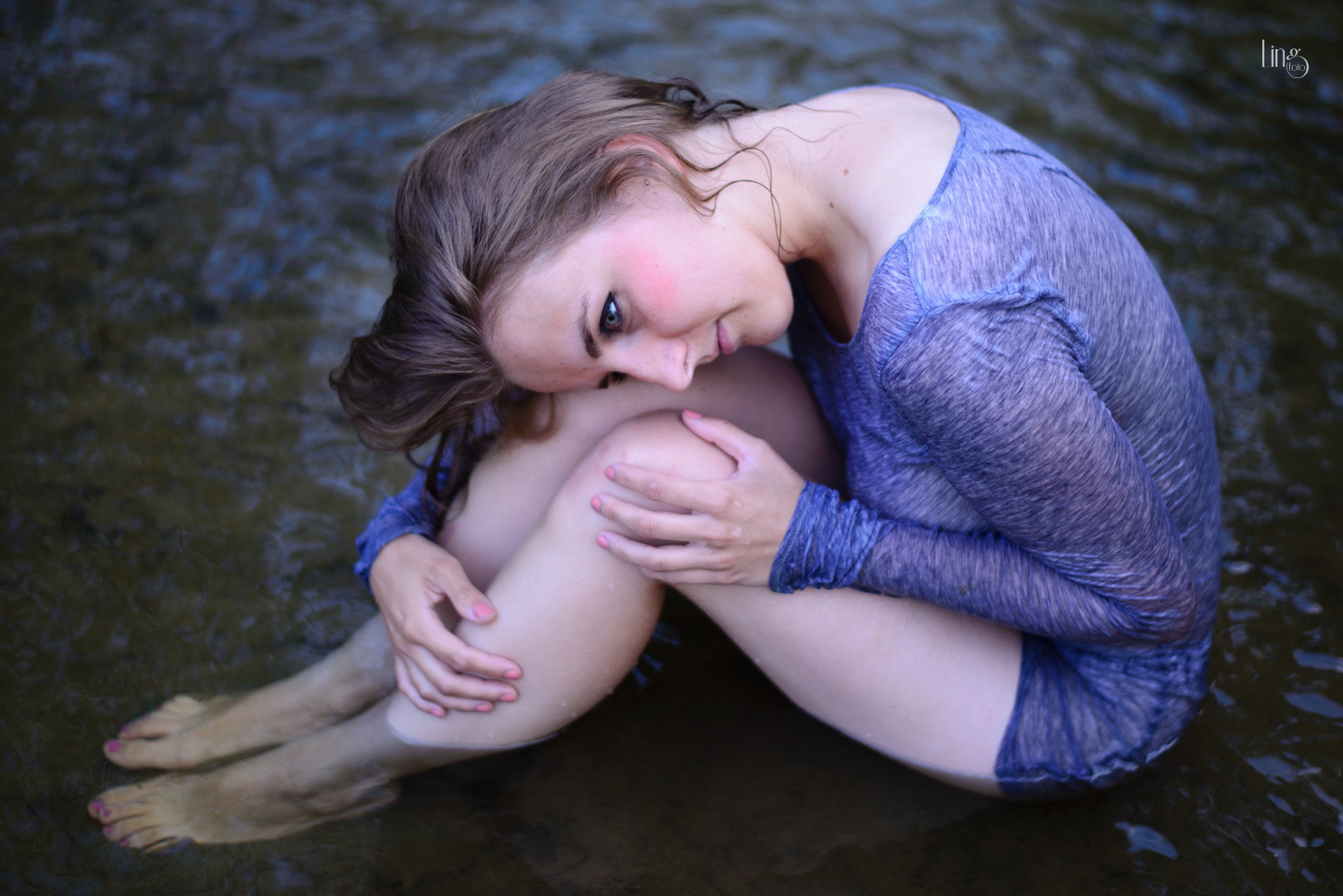 Młoda kobieta w mokrym, błękitnym ubraniu siedzi w wodzie, obejmując kolana i spoglądając w dół, z mokrymi włosami opadającymi na twarz.