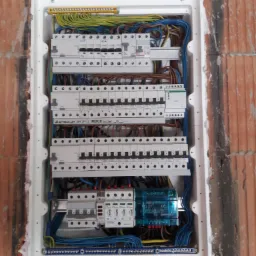 Rozdzielnica elektryczna w trakcie montażu, widoczne kable, bezpieczniki, listwy zaciskowe i obudowa zamontowana w ścianie z cegły. Część przewodów oznaczona opisami. Widoczne zabezpieczenia...