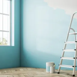 Pokój w trakcie malowania na turkusowo, z widoczną białą drabiną, puszkami farby i fragmentem ściany pokrytym białą farbą, drewniana podłoga, okno z widokiem na palmy.