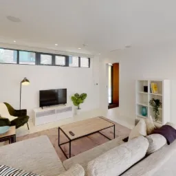 Jasne wnętrze salonu z białymi ścianami, drewnianą podłogą i nowoczesnymi meblami, w tym szarą sofą, zielonym fotelem i stolikiem kawowym na metalowej ramie.